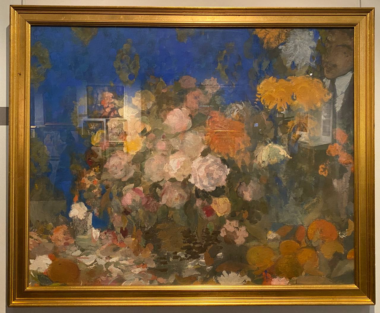 Painting in a gold frame: a vivid bouquet of peonies and chrysanthemums on a blue background, oranges below, a man’s profile in the upper right corner.