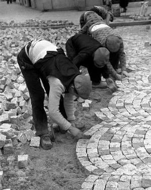 Black-and-white photograph of three workers bent over the ground, laying cobblestones in arcs, with piles of stones beside them.