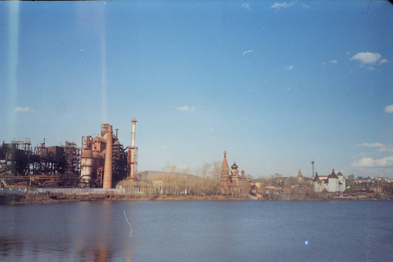 Au bord d'un lac, des bâtiments industriels en brique avec cheminées et structures de hauts fourneaux, et à droite une église à coupoles et des bâtiments blancs.
