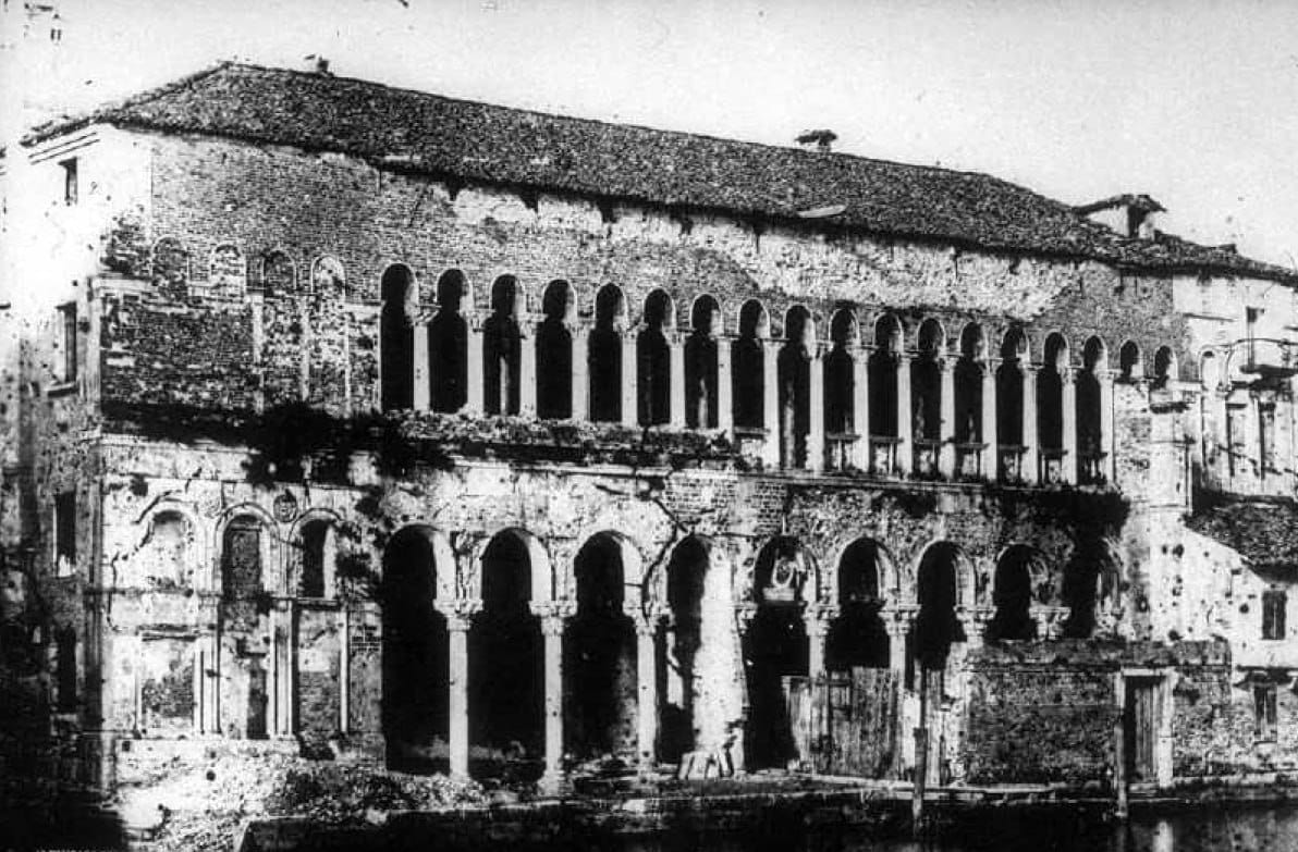 Photo en noir et blanc d'un édifice en brique délabré, avec deux niveaux de galeries à arcades sur de fines colonnes et un toit de tuiles. De l'eau sombre devant la façade.