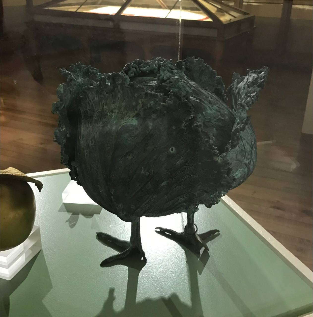 A sculpture shaped like a cabbage head on two bird legs stands in a museum display case on a green table.