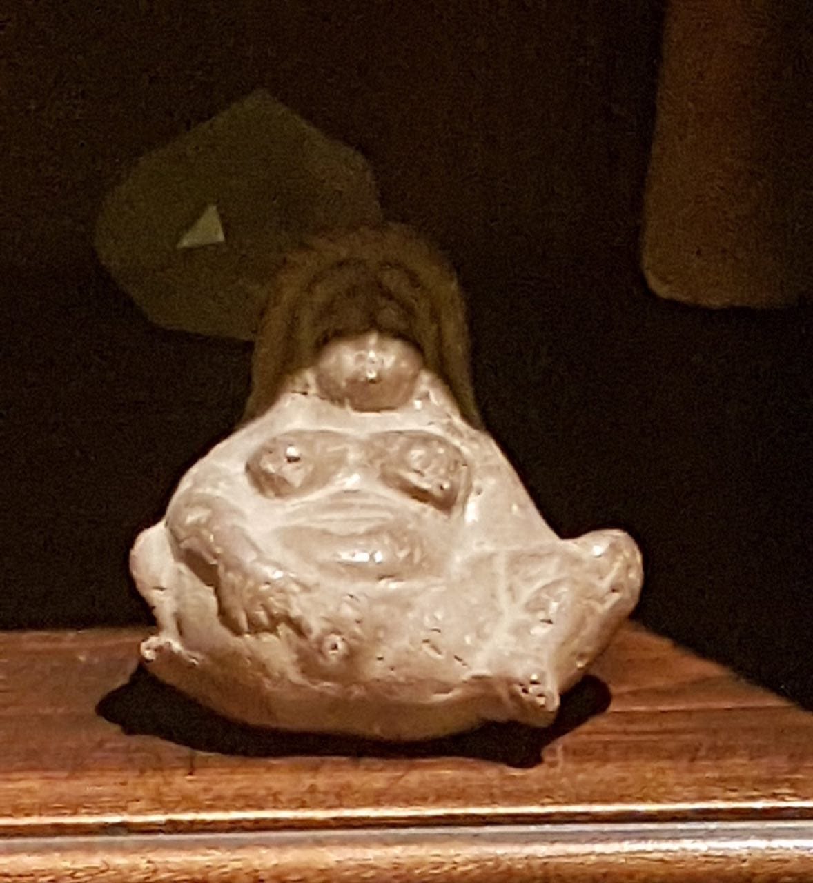 A small pale figurine of Baubo with a tuft of hair on its head stands on a wooden shelf against a dark background.