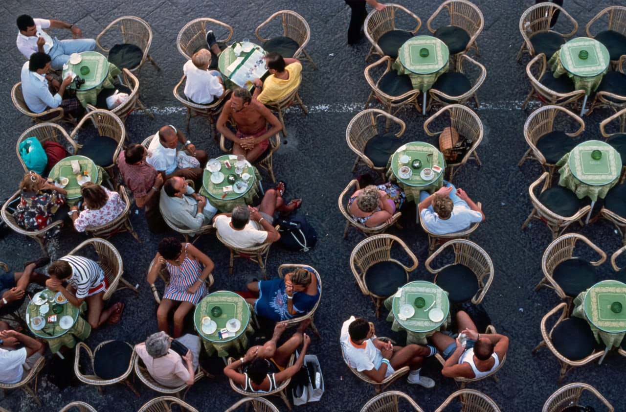 Overhead view of people seated at round tables with green tablecloths and wicker chairs on an open-air café terrace.