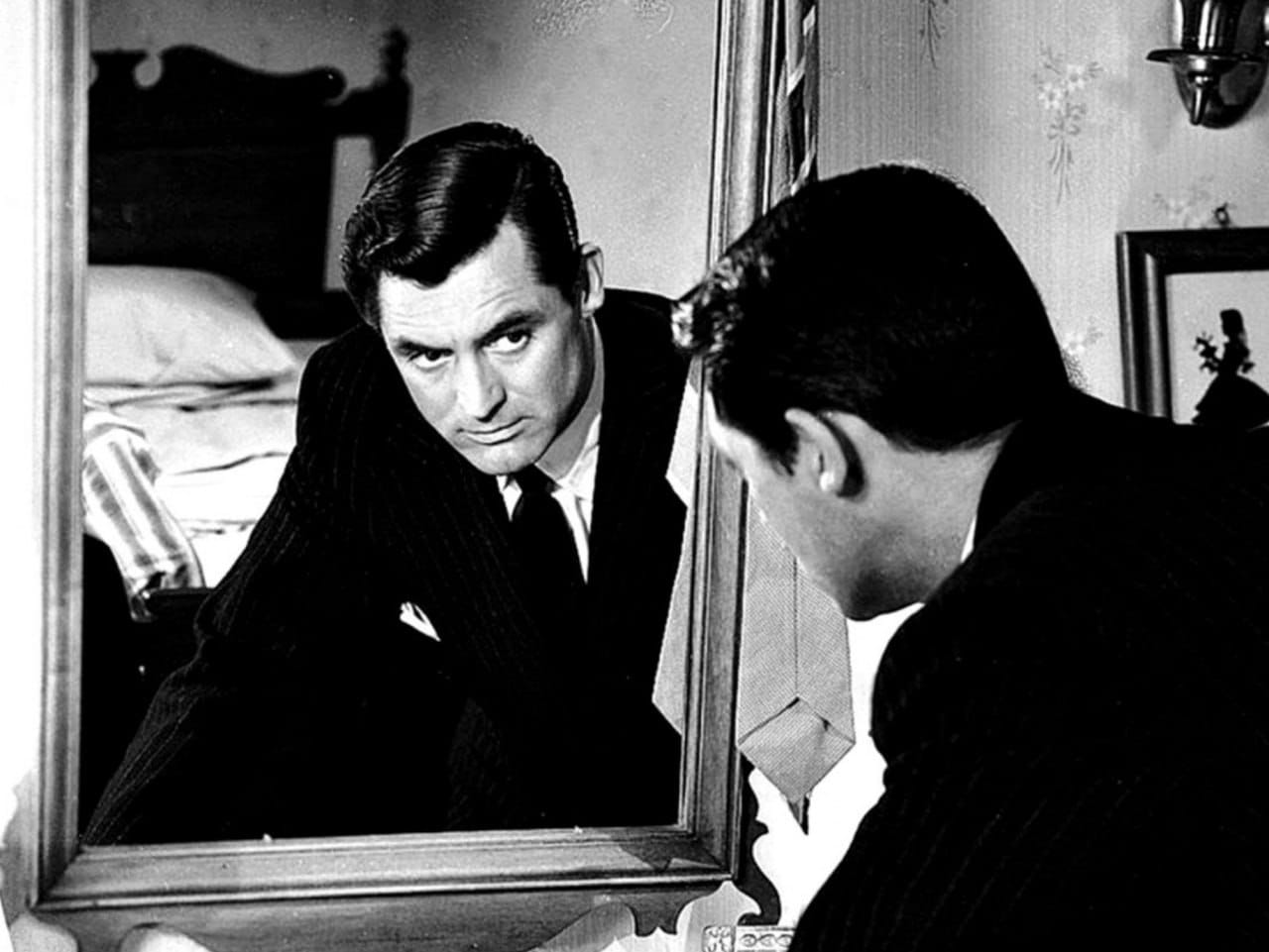 Black-and-white still of a man in a dark pinstriped suit looking at his reflection in a mirror in a bedroom.