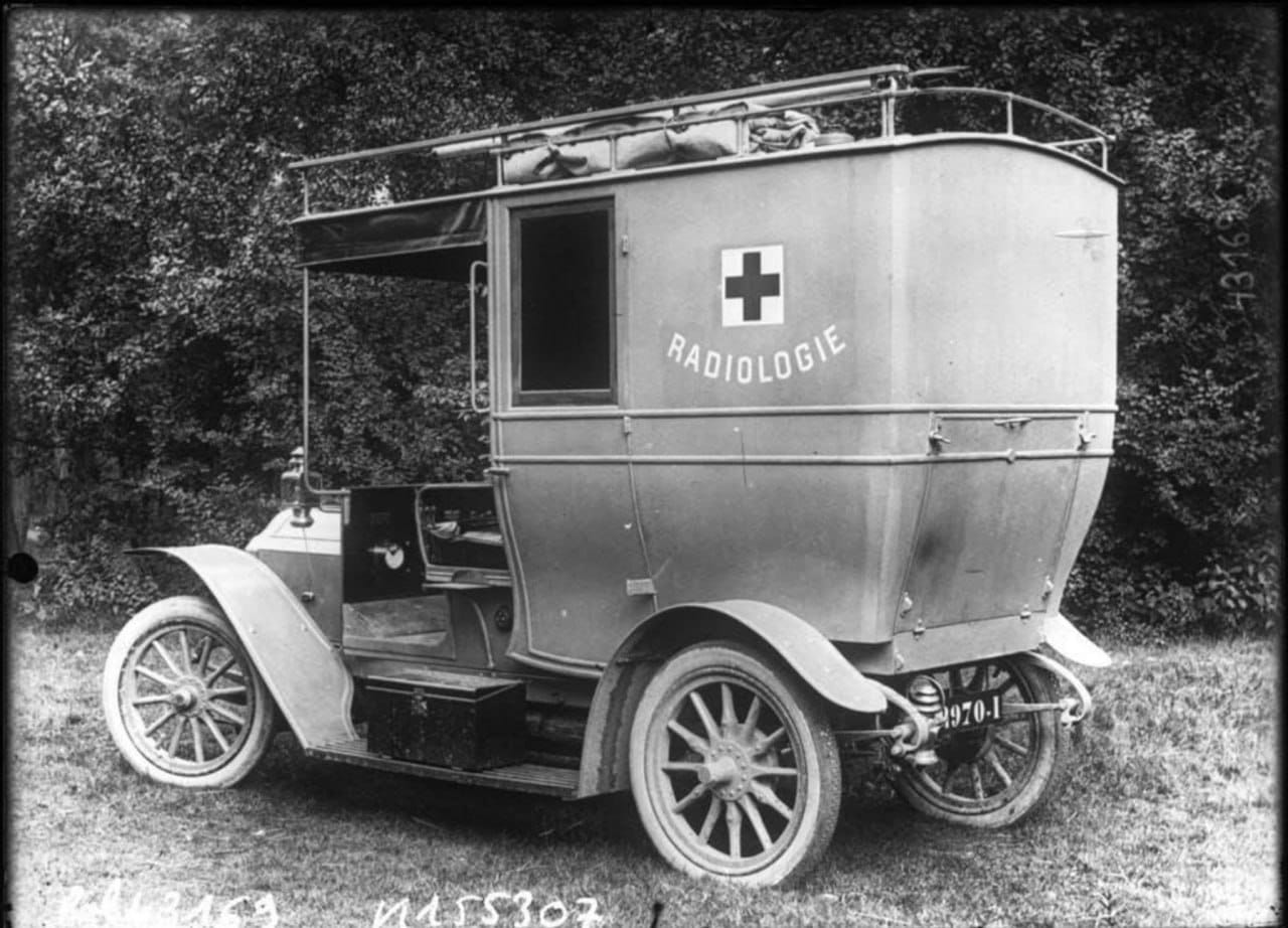 An old car with a tall body, a cross and the word “Radiologie” on its side stands on grass in front of trees.