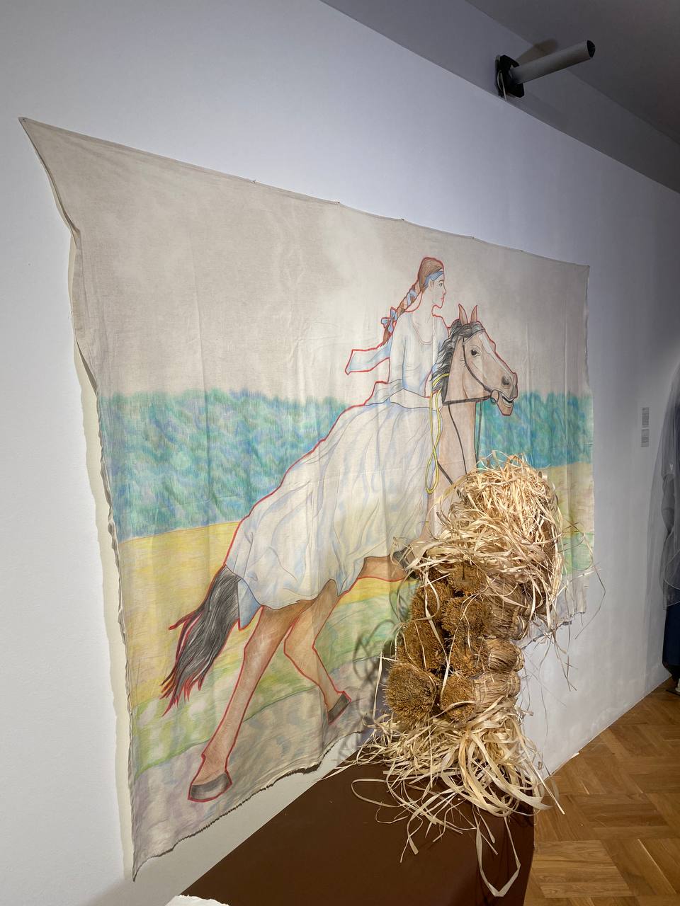 Une toile accrochée au mur montre une jeune femme tressée en robe blanche galopant à cheval, en couleurs. Devant, une sculpture de paille et de fibres sur un socle brun.