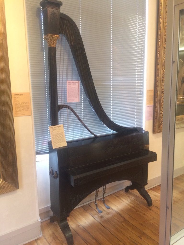 Un claviharpe noir dans une salle de musée, avec un cadre en forme de harpe posé sur un caisson à clavier et pédales.