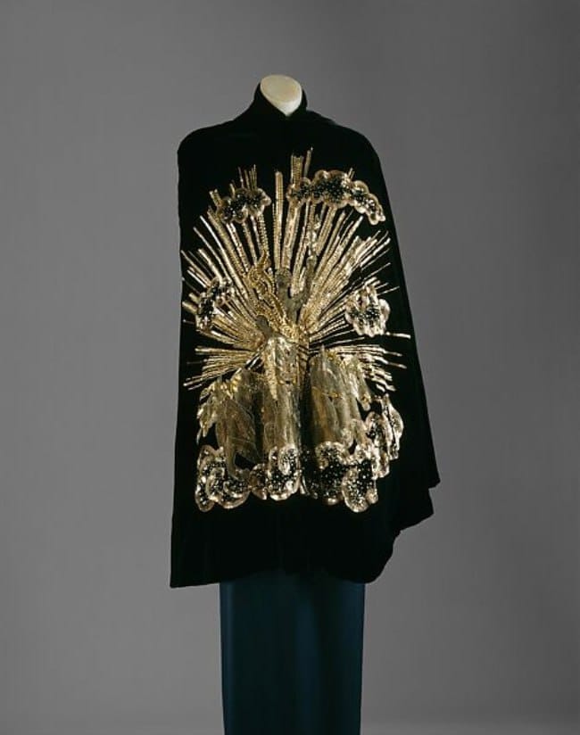 Cape en velours noir sur un mannequin, brodée d'or dans le dos : une figure au milieu de rayons, entourée de nuages perlés. Une robe bleu foncé dépasse en dessous.