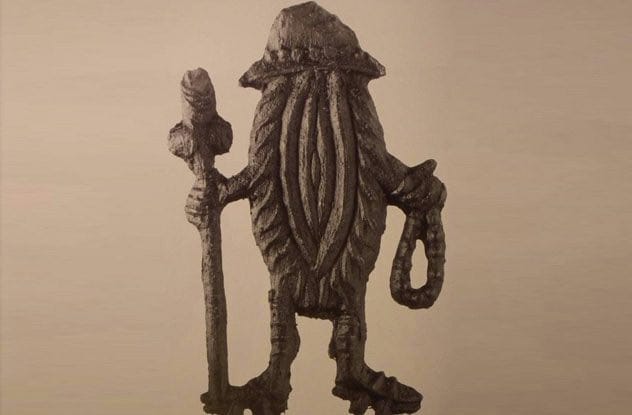 Dark metal badge showing a figure in a hat with a staff in one hand and a rope in the other, its body shaped like a vagina.