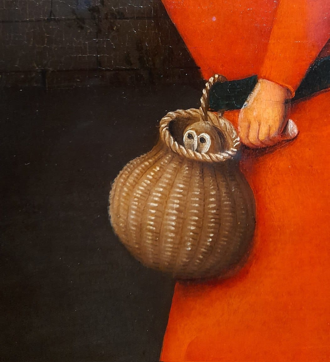 Détail de peinture : une main dans une manche rouge tient un panier d'osier d'où dépasse une petite chouette. Le fond est sombre, la peinture craquelée.