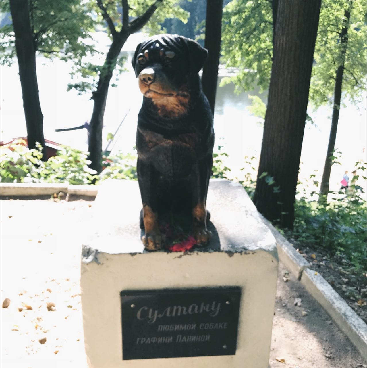 Bronze sculpture of a seated Rottweiler on a white pedestal with a plaque reading “To Sultan, beloved dog of Countess Panina”, with trees and water behind.