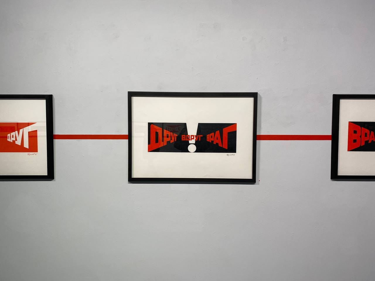 Trois estampes graphiques sous cadre noir sur un mur clair, avec des lettres rouges et noires : «Друг», «Вдруг», «Враг». Une ligne rouge horizontale relie les cadres.