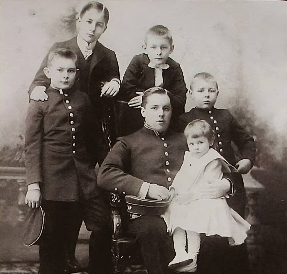 Ancienne photographie en noir et blanc : un jeune homme en uniforme boutonné est assis, une fillette en robe claire sur les genoux, entouré de quatre garçons debout.