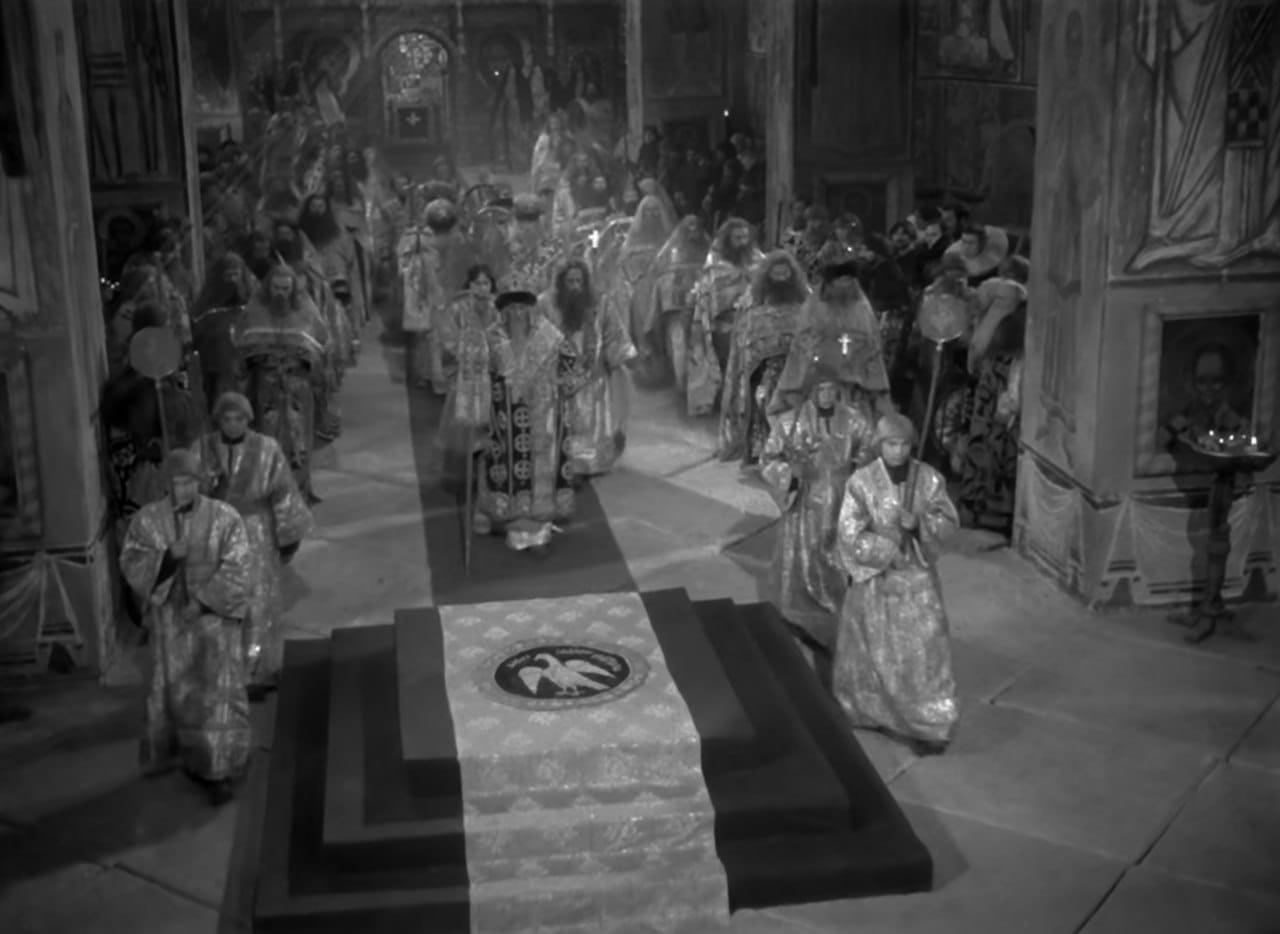 Image en noir et blanc : des ecclésiastiques en habits brodés avancent sur un tapis dans une église à fresques vers une estrade ornée d'un aigle.