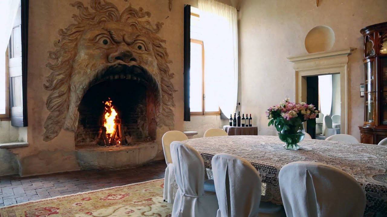 Un feu brûle dans une cheminée sculptée en visage grimaçant à la chevelure ondulée. À droite, une table en dentelle, des chaises housses blanches, un bouquet et des bouteilles.