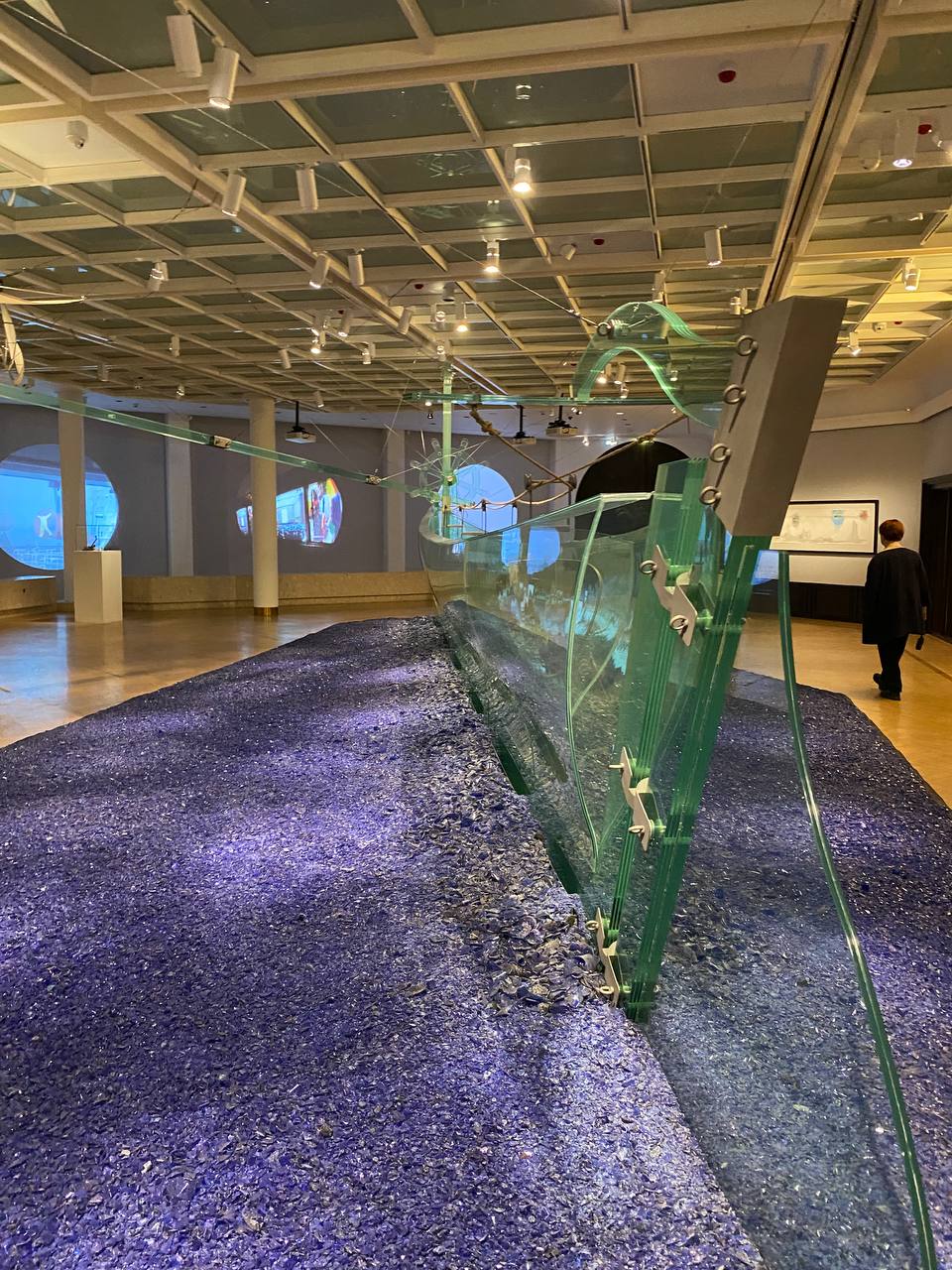 Dans une salle de musée, un long lit d'éclats de verre bleu porte des panneaux de verre courbes fixés par des attaches métalliques. Des projections rondes ornent les murs ; un visiteur passe