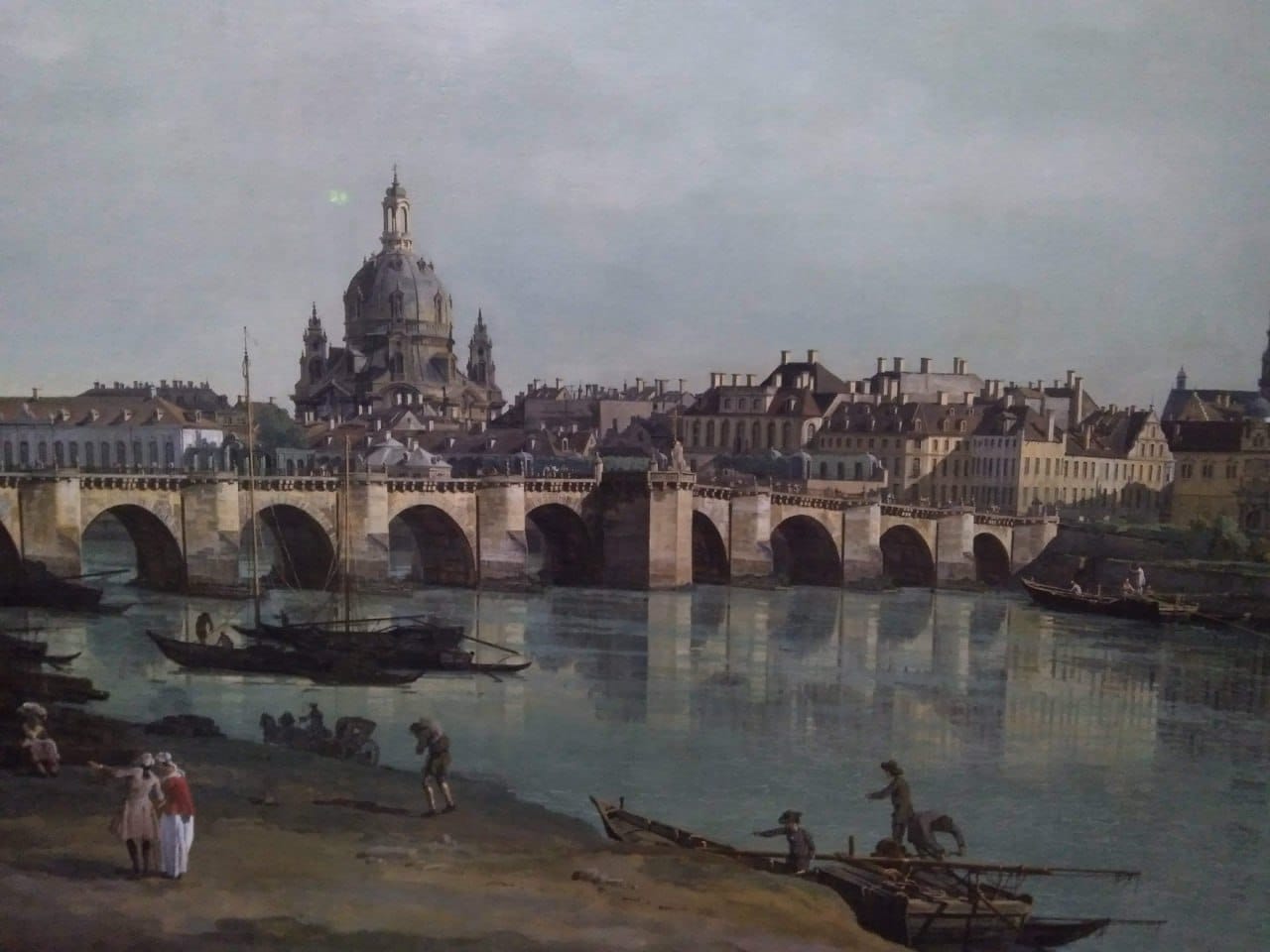 Tableau d'un large fleuve traversé par un pont de pierre à arches, avec des maisons et un grand dôme sous un ciel couvert. Sur la rive, des gens en costume du XVIIIe siècle et des bateaux.