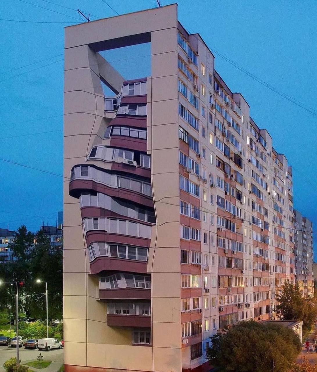 End wall of a high-rise residential block painted with warped balconies that create the illusion of a bending façade, seen against the evening sky.