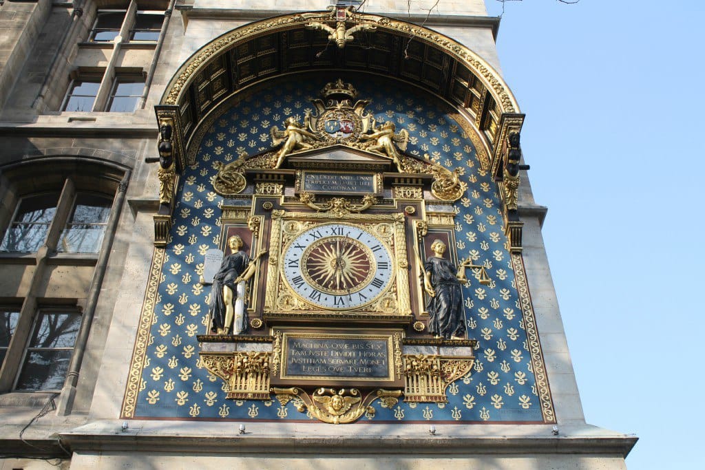 Horloge de tour dorée aux chiffres romains, avec un soleil au centre du cadran, flanquée de deux statues, sur un fond bleu semé de fleurs de lys dorées.