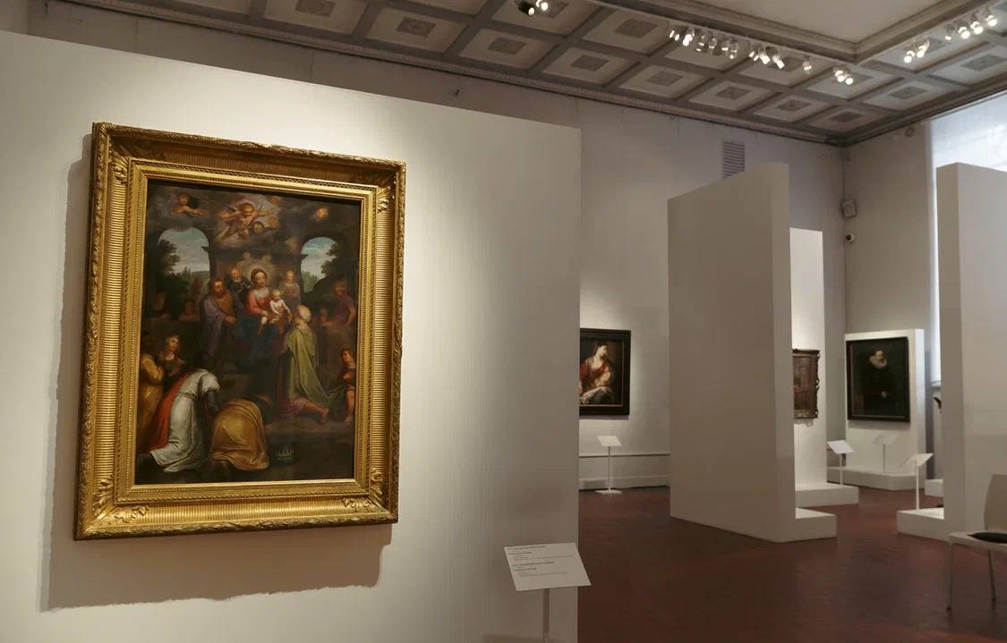Dans une salle de musée, un ancien tableau de la Vierge, de l’Enfant et des mages agenouillés, au cadre doré, orne un mur blanc. D’autres tableaux et cloisons se voient au fond.