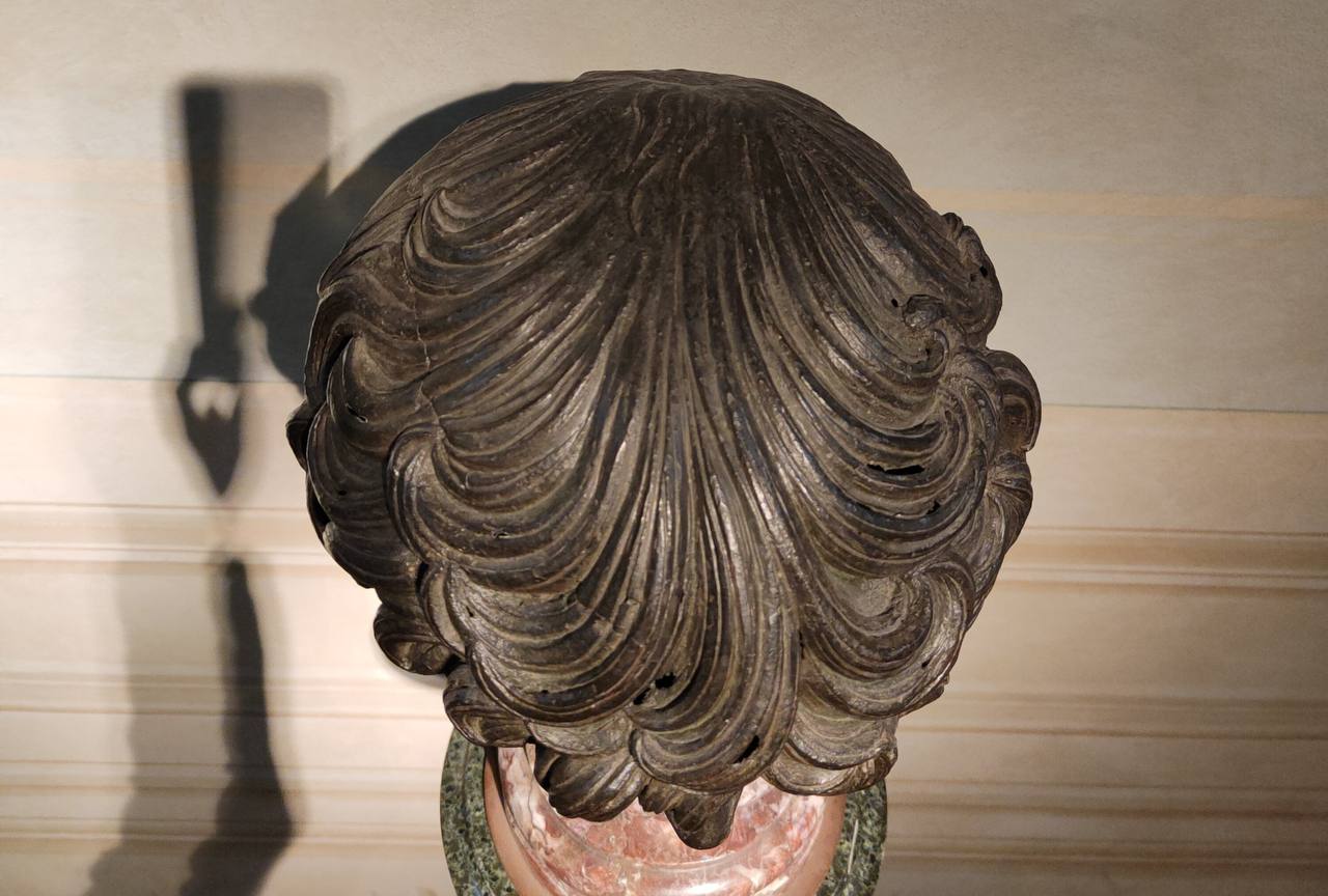 Tête sculptée sombre vue de dessus, aux cheveux taillés en mèches ondulées. Elle repose sur un socle de marbre rose et vert, une ombre se projetant sur le mur.