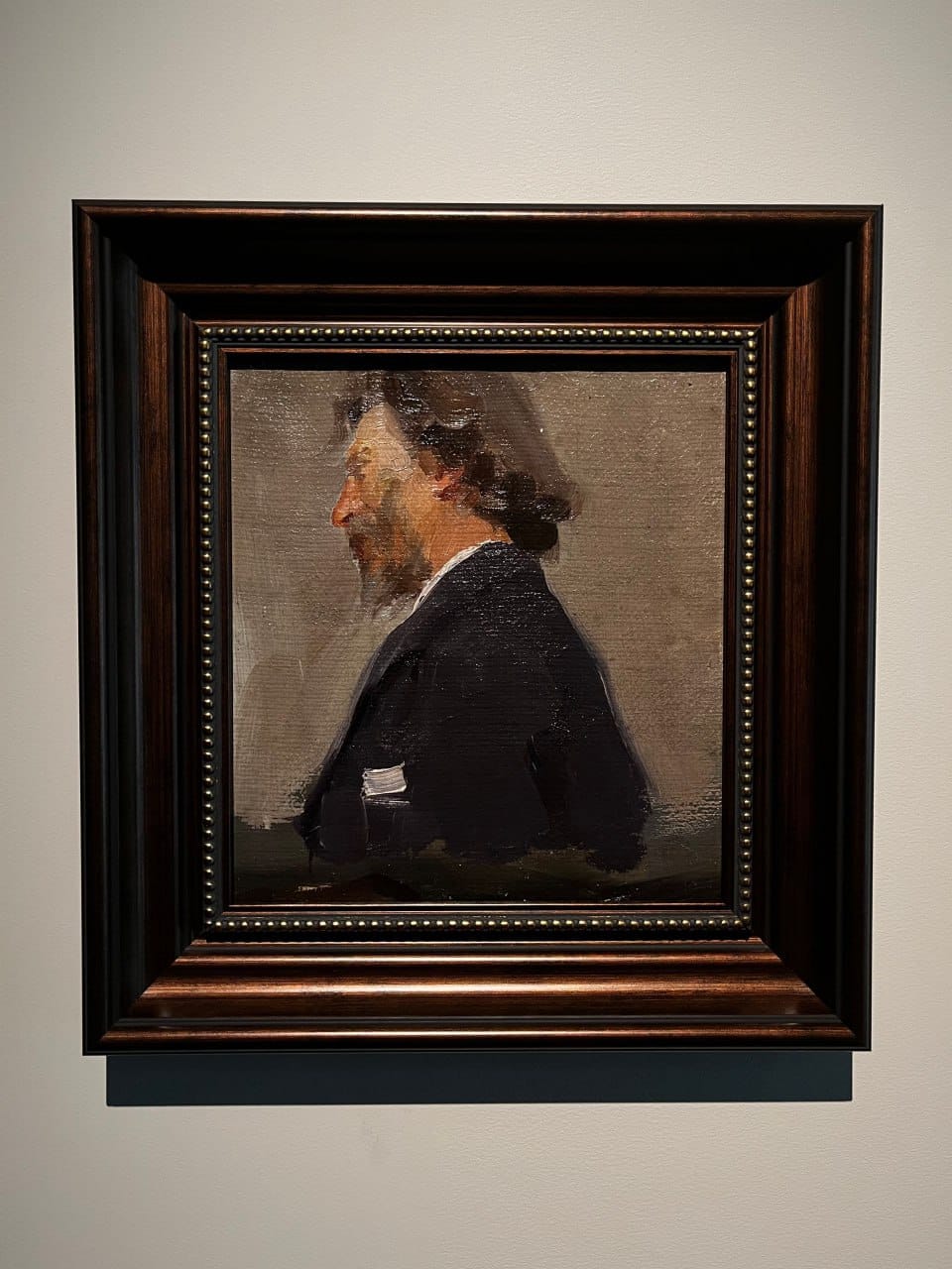 Étude à l'huile d'un homme barbu aux longs cheveux sombres, de profil vers la gauche, en habit sombre sur fond gris. Elle est présentée dans un large cadre brun sur un mur clair.