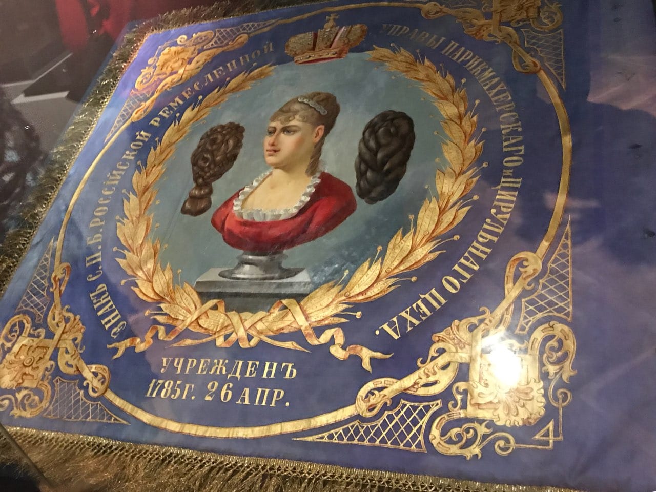 Алтын шашақты көк жібек ту, лавр шоқтығы және ортасында қызыл көйлекті, биік шаштүзетілімді әйел бюсті мен екі жағындағы екі парик.