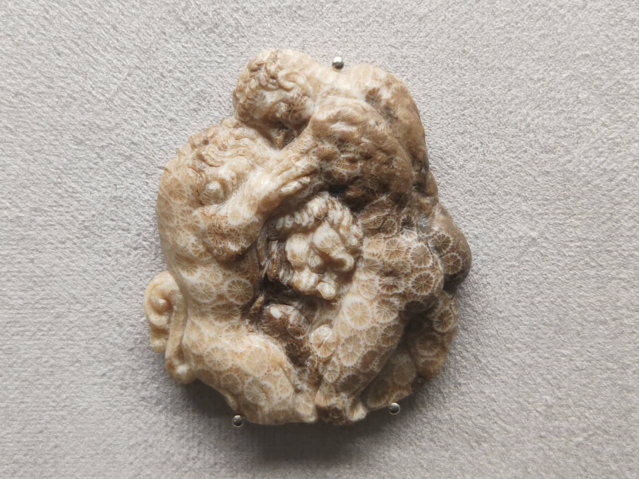 Carved relief in pale stone with a fossil coral pattern showing a nude figure wrestling a lion, held by small prongs against a grey background.