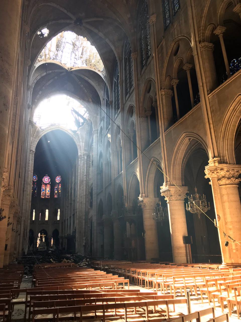 Dans une cathédrale gothique, des rayons de soleil traversent un trou de la voûte et éclairent colonnes et rangées de chaises en bois ; au fond, vitraux et gravats.