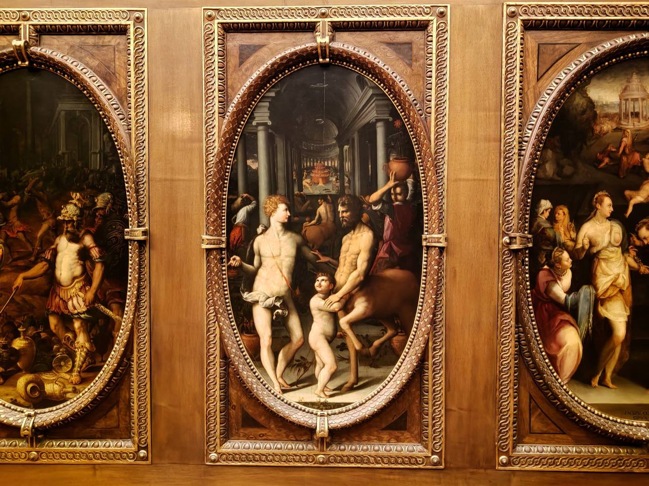 Trois tableaux ovales dans des cadres dorés sur un mur lambrissé. Au centre, un jeune homme nu avec un arc, un centaure et un enfant devant une salle à colonnes.