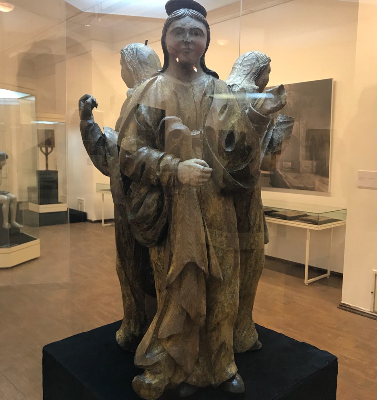 Groupe sculpté de trois anges en bois, dont celui du centre porte un nimbe, exposé sous verre dans une salle de musée.