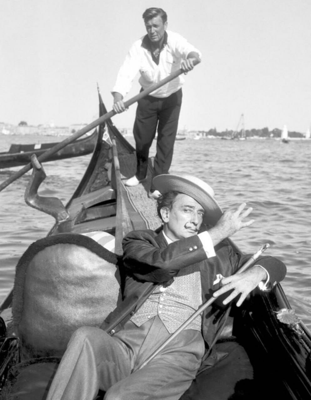 Photo en noir et blanc : un homme moustachu en canotier et gilet, à demi allongé dans une gondole, la main levée. Derrière lui, un gondolier à la rame ; l'eau au loin.
