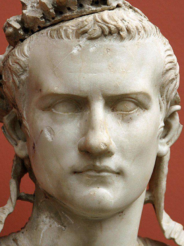 Gros plan sur une tête d'homme en marbre aux mèches courtes ramenées sur le front, avec une couronne abîmée et des rubans près des oreilles. Yeux sans pupilles, fond rouge.