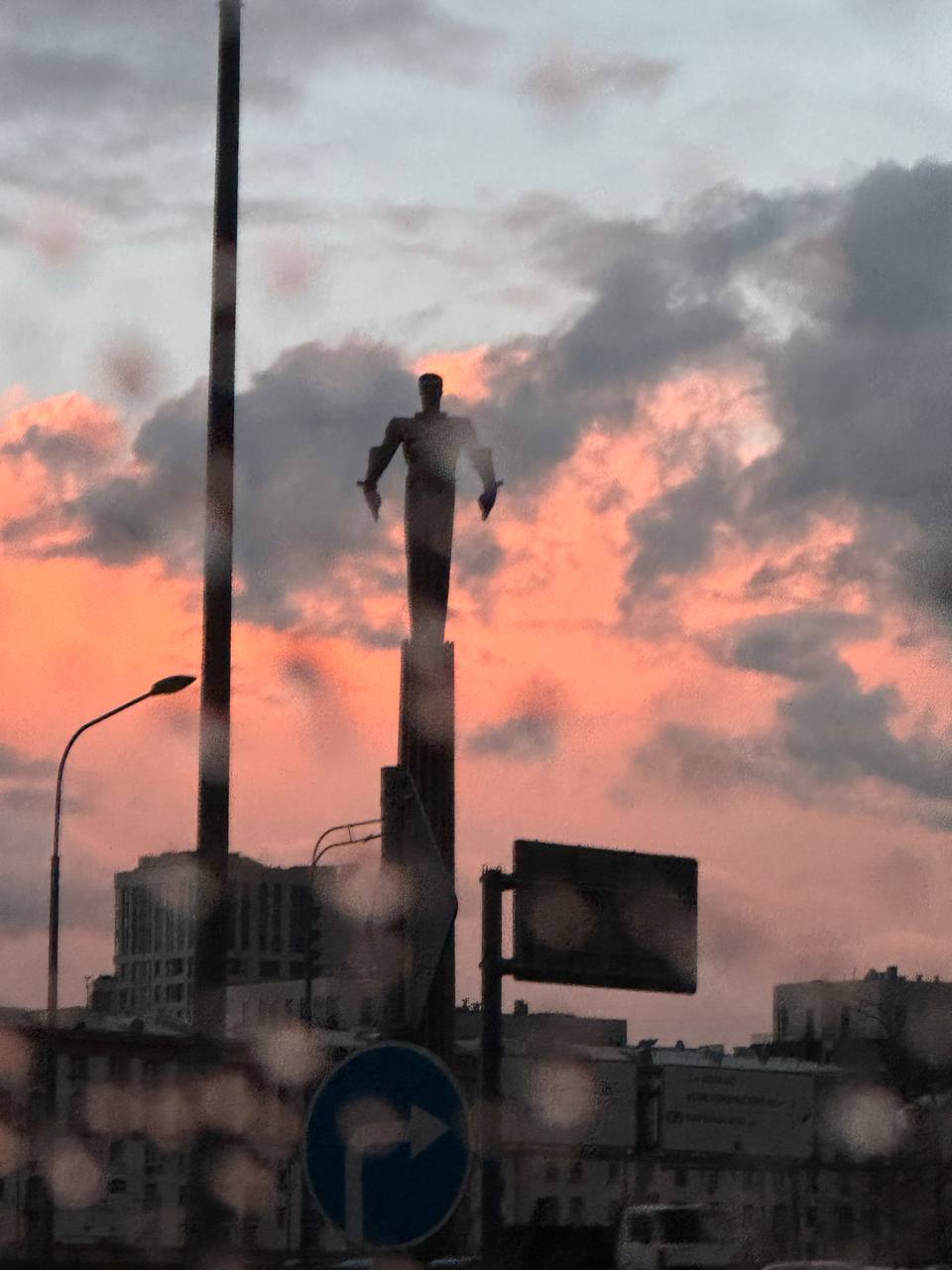 Silhouette d'une statue au sommet d'une haute colonne devant des nuages roses et gris, prise à travers une vitre mouillée, avec un panneau routier et des immeubles en bas.