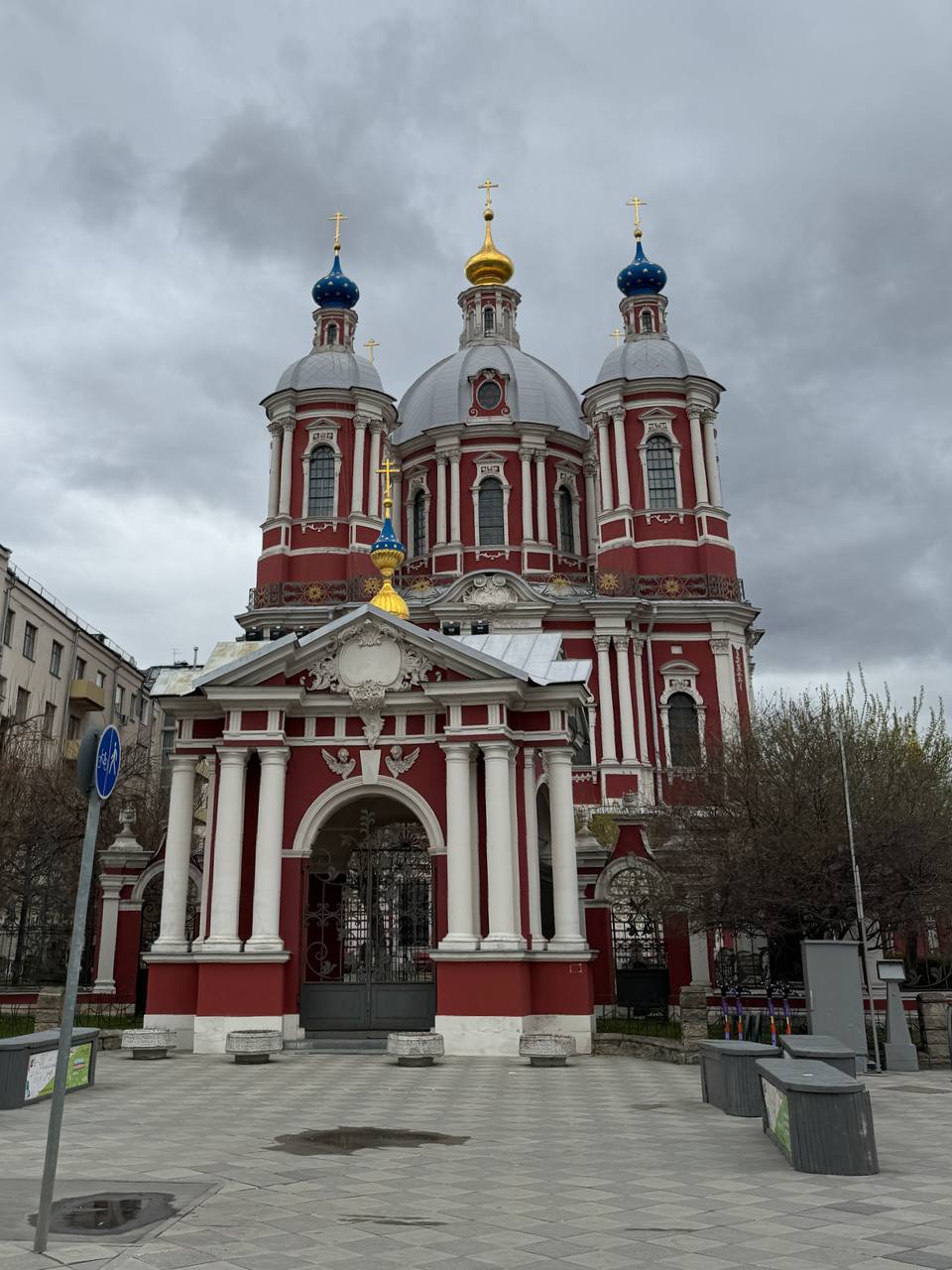A red church with white columns, one golden dome and blue domes with stars, under an overcast sky.