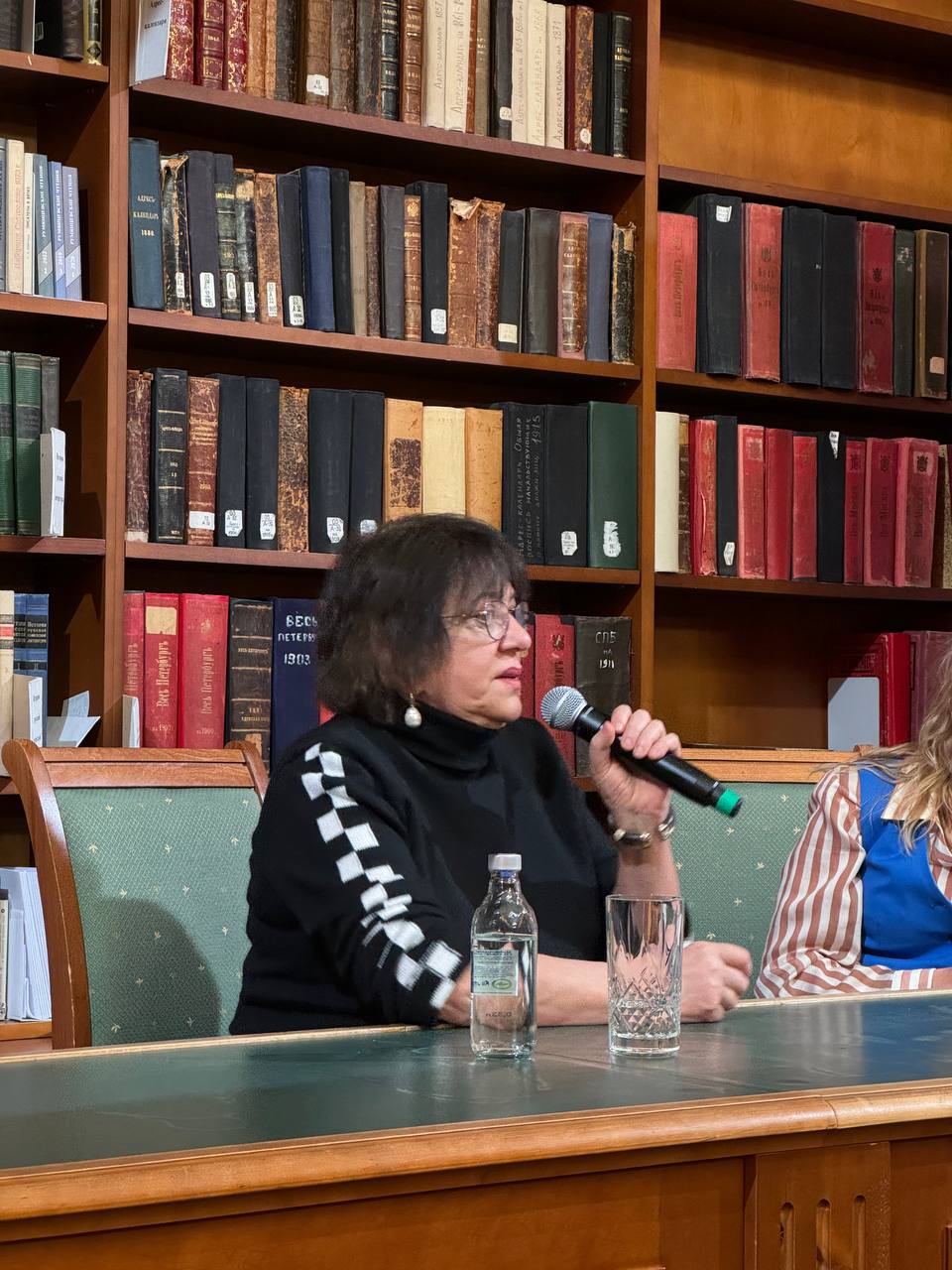 Une femme en pull noir et lunettes parle dans un micro à une table, avec une bouteille d'eau et un verre. Derrière elle, des étagères d'anciens livres, dont Tout Pétersbourg et des annuaires