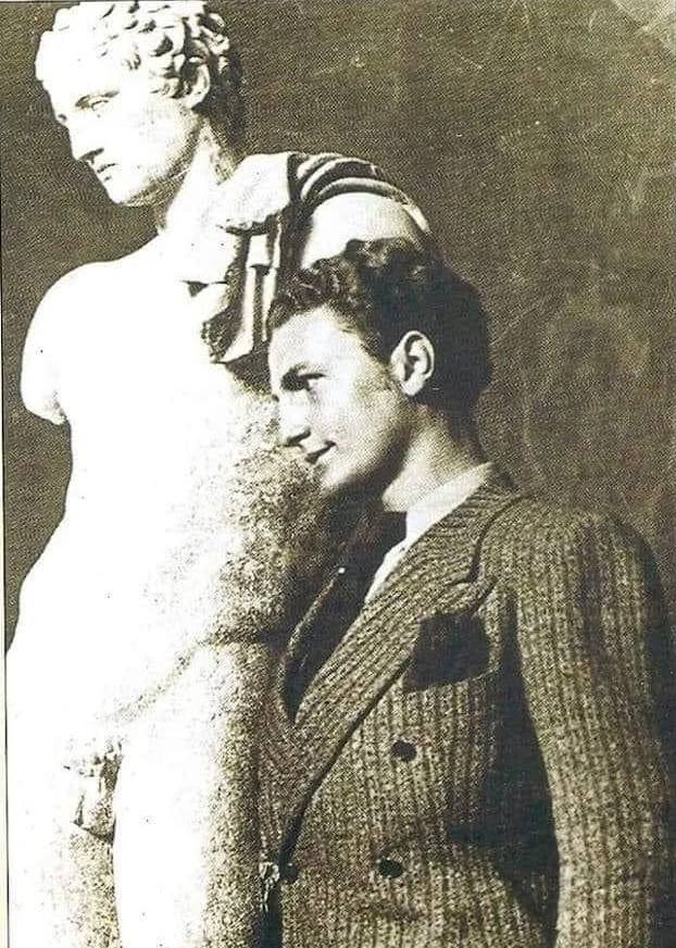 Black-and-white photograph of a young man in a jacket standing in profile beside a marble statue of Hermes.