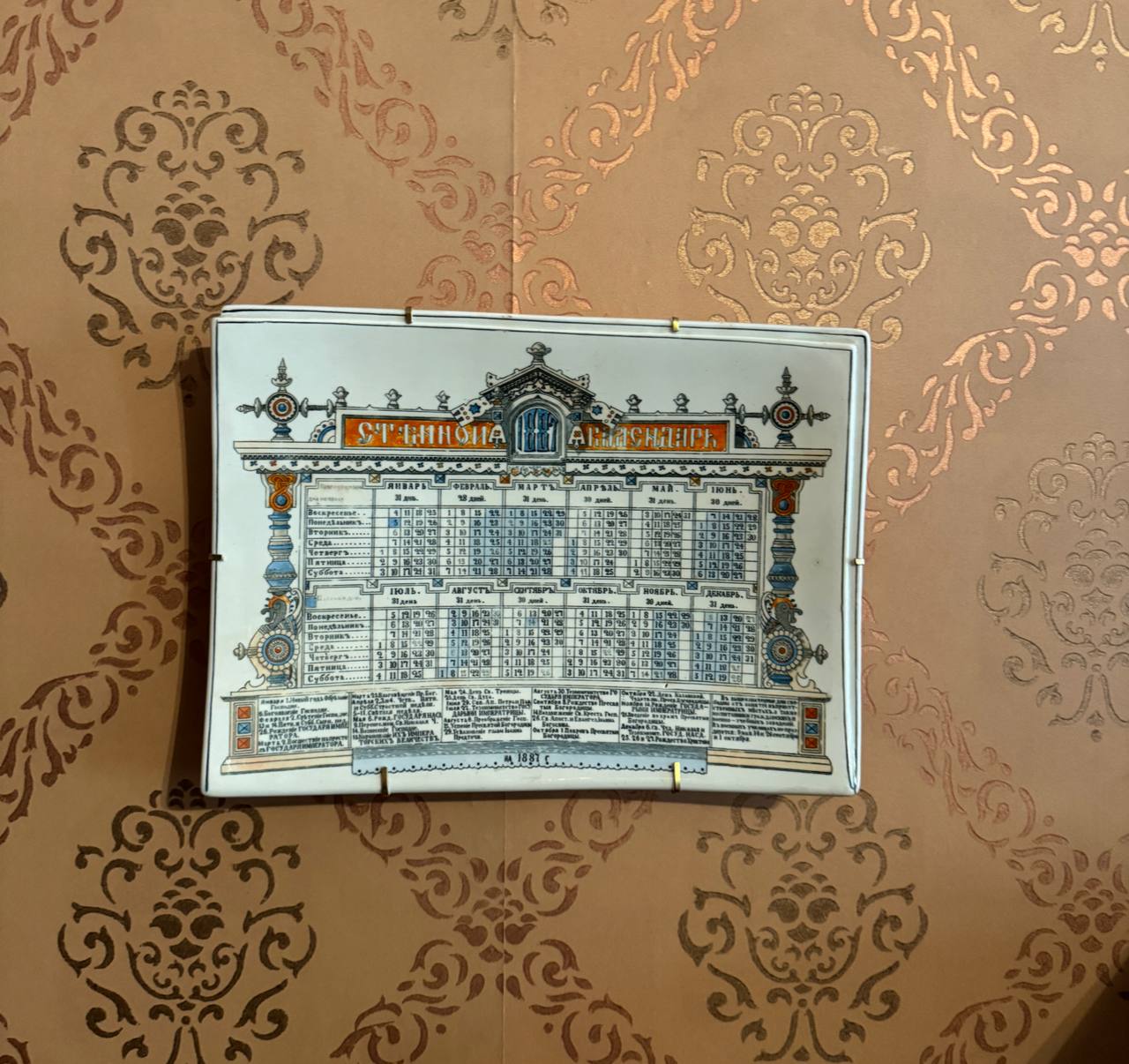 A rectangular ceramic dish printed with a calendar for 1887 hangs on a wall with copper-coloured patterned wallpaper.