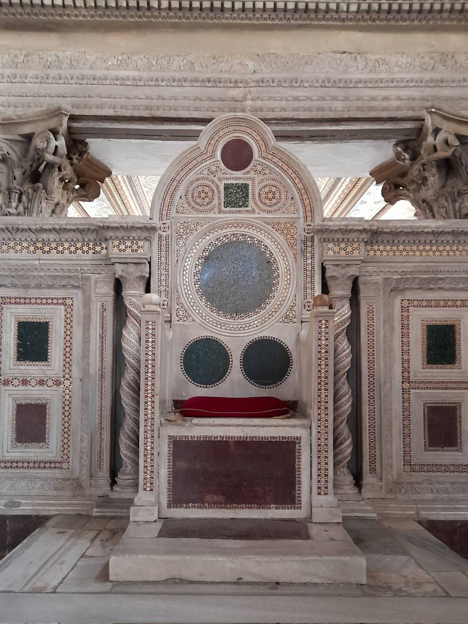Cathèdre en marbre ornée de mosaïques, de médaillons de pierre verte et rouge, de colonnettes torses et d'un coussin rouge sur l'assise.
