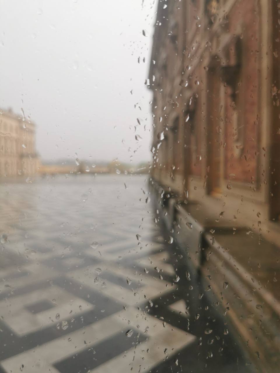 Vue à travers une vitre mouillée sur une cour au dallage géométrique noir et blanc. À droite, une façade de brique et de pierre ; au loin, une aile floue dans la brume.