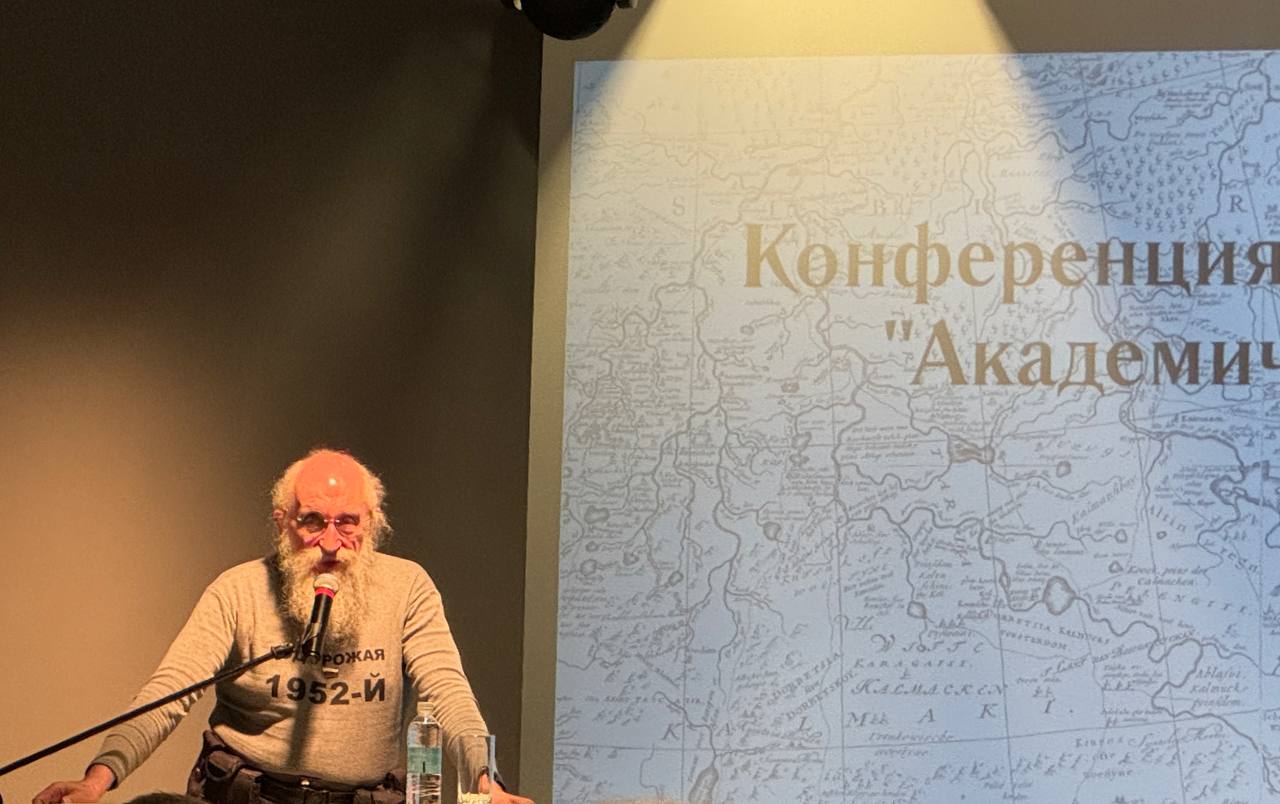 Көзілдірікті, сақалды, сұр жемпірлі ер адам «Конференция» жазуы мен ескі карта көрсетілген экран алдында микрофонға сөйлеп тұр.