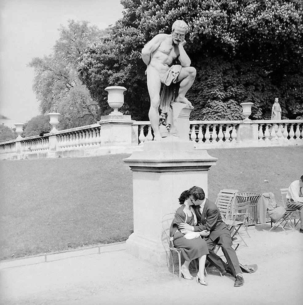 Black-and-white photo of a man and a woman kissing on a chair in a park beneath a statue of a pensive nude bearded man on a pedestal.
