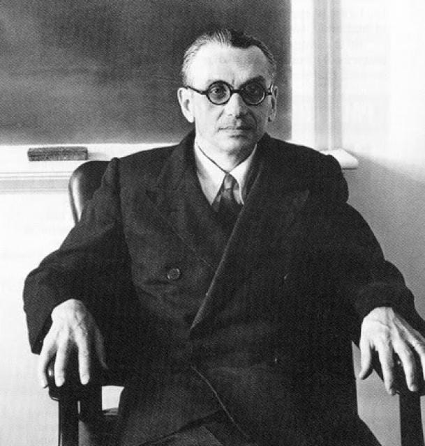 Photographie en noir et blanc d'un homme à lunettes rondes et en costume sombre, assis dans un fauteuil devant un tableau noir.