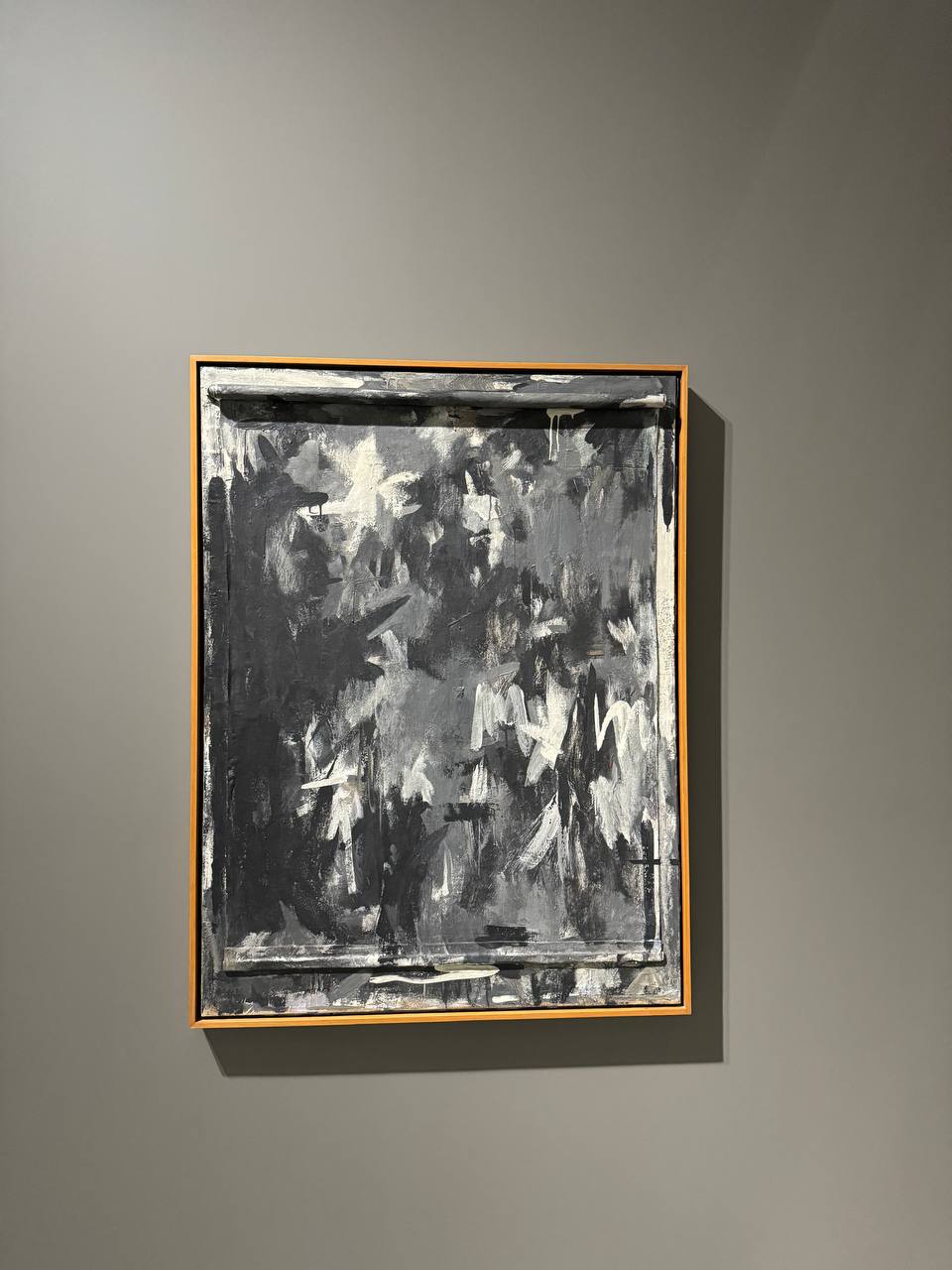 Toile abstraite aux coups de pinceau noirs, gris et blancs, dans un cadre en bois clair sur un mur gris foncé. Le haut et le bas de la toile sont enroulés comme un rouleau.