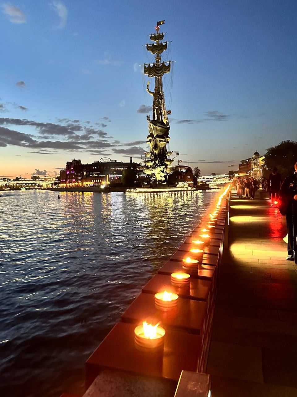 Une rangée de bougies allumées sur le parapet du quai, des passants à droite. Au loin, au-dessus du fleuve, un monument en forme de voilier se dresse dans le ciel du soir.