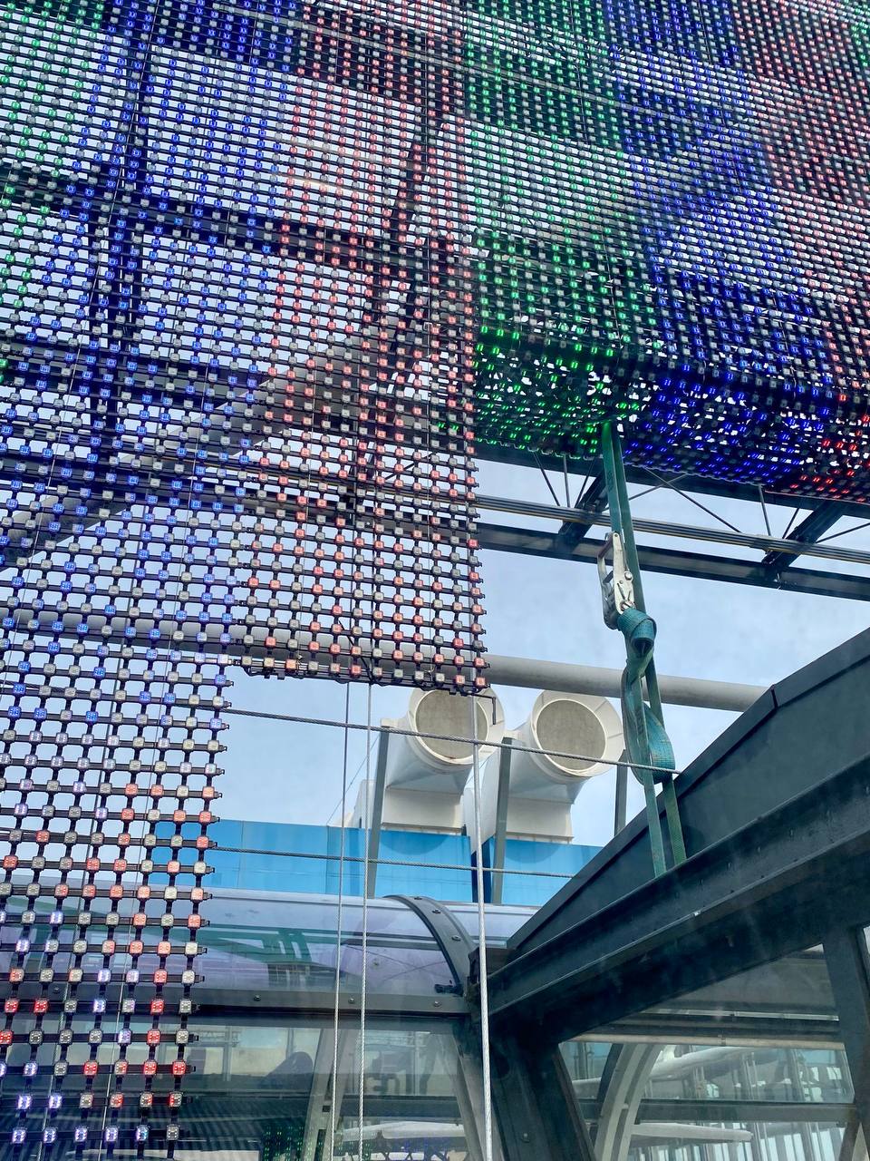 A mesh LED panel with blue, red and green lights on a facade, seen from below, with white ventilation pipes and glass structures behind.