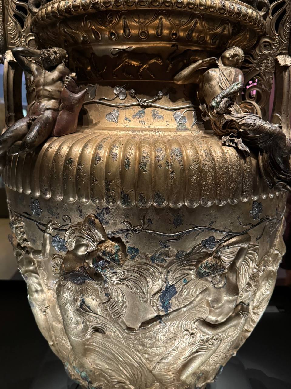 Gros plan d'un vase ancien en bronze doré : épaule côtelée, feuilles de vigne en argent, figures allongées près du col et relief de figures féminines sur la panse, avec patine.