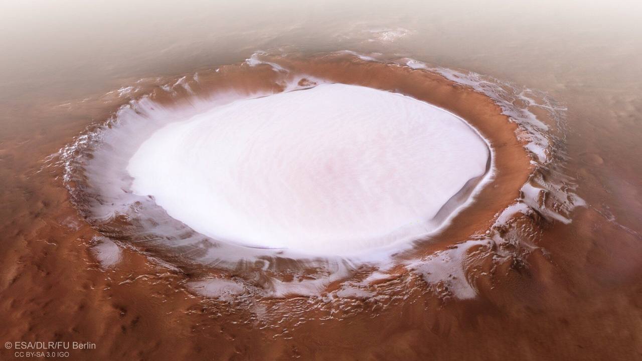 Cratère circulaire aux bords roux, au fond couvert de glace blanche, entouré d'un terrain désertique brun-orangé dans une légère brume.