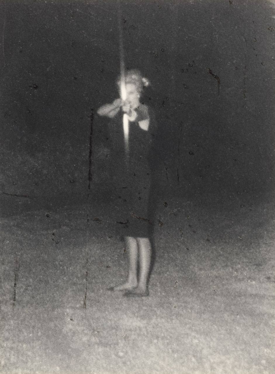 Blurred black-and-white night photograph of a woman in dark clothing standing barefoot on grass and aiming a bow.