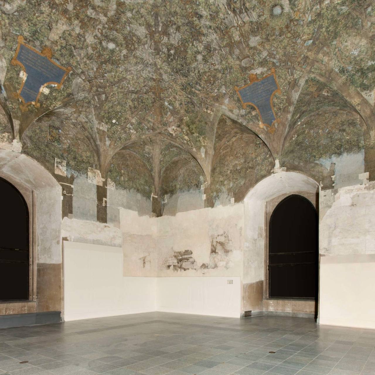 Salle voûtée au plafond peint de branches et de feuillages, avec deux cartouches bleus à inscriptions. Murs en partie dégradés, deux arcades sombres, sol dallé.